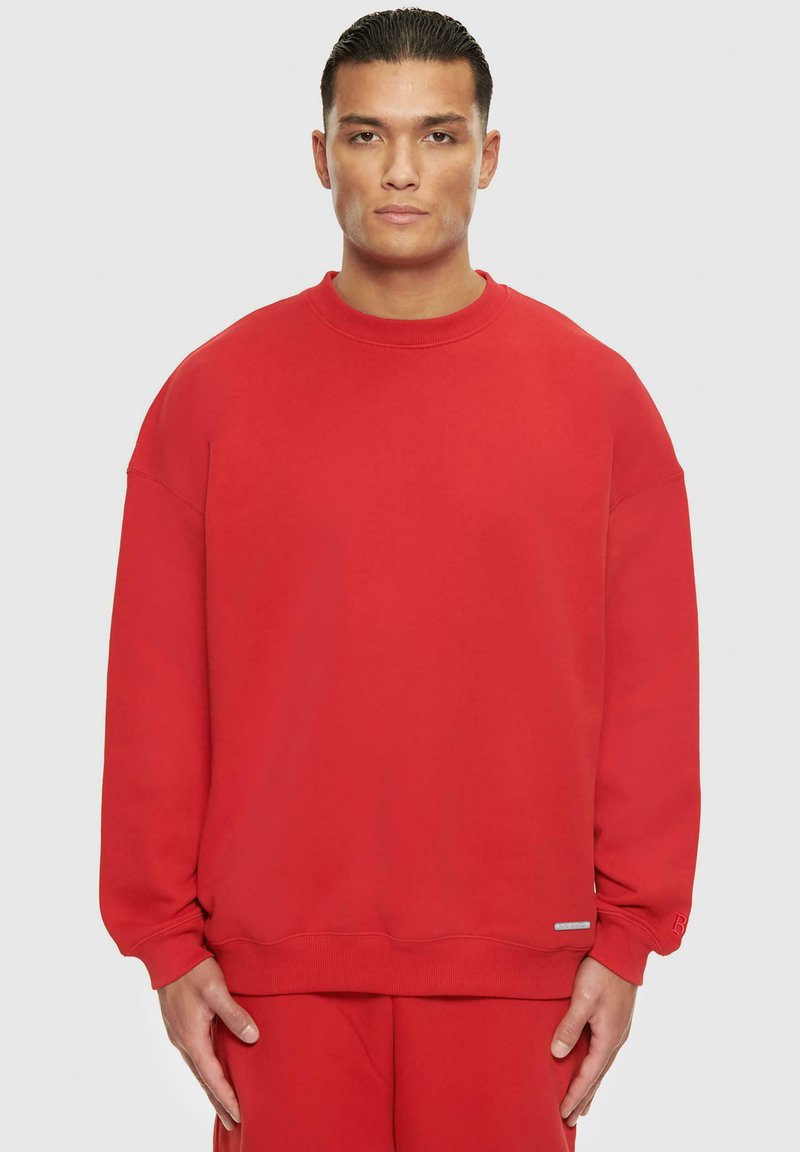 Rotes Sweatshirt mit rundem Halsausschnitt, lockerer Passform, gerippten Bündchen und Saum. Weicher Stoff und minimaler Markenaufdruck auf der unteren linken Seite.