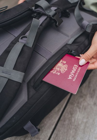 Zaino in tessuto nero con spallacci grigi; un passaporto spagnolo rosso è parzialmente visibile in una tasca con zip. La superficie è texture e liscia.