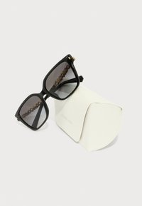 Čierne oversized slnečné okuliare so šedými gradientnými sklami. Sú vybavené zlatým akcentom na ráme a odpočívajú na mäkkom bielom puzdre s označením "Michael Kors."