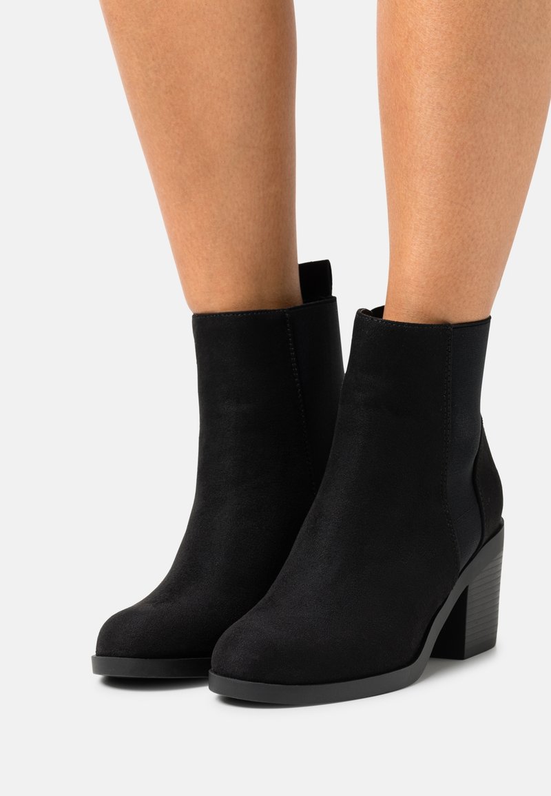 Bottines noires en daim, avec un bout arrondi, des panneaux latéraux élastiques et un talon en bloc texturé. Surface lisse avec des accents minimaux.