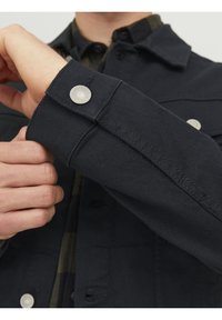 Man som fäster en silverknapp på ärmen till en mörkblå denimjacka, bärandes en mörkrutig skjorta under.