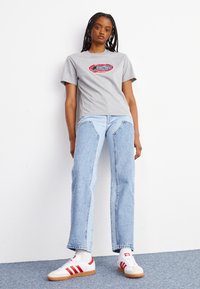 T-shirt en coton gris avec un imprimé graphique coloré, associé à un jean taille haute bleu clair avec des détails de coutures uniques, et des baskets blanches à rayures rouges.