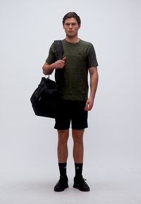 Νεαρός άνδρας που φοράει πράσινο t-shirt Adidas, μαύρα σορτς και μαύρα αθλητικά παπούτσια, κρατώντας μια μαύρη τσάντα γυμναστηρίου στον ώμο του.