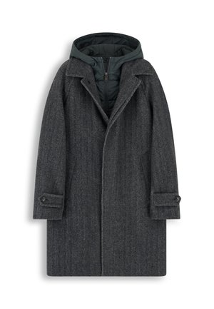 Un homme porte une tenue grise en couches avec un manteau en laine, un cardigan et un pantalon, assortie à des baskets noires sur un fond blanc uni.