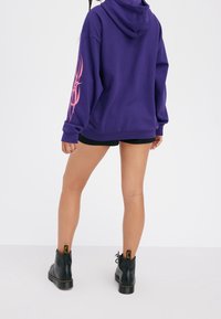 Felpa viola con una grafica rosa sulla manica sinistra, indossata con pantaloni corti neri e stivaletti neri alla caviglia. Materiale morbido, vestibilità over.