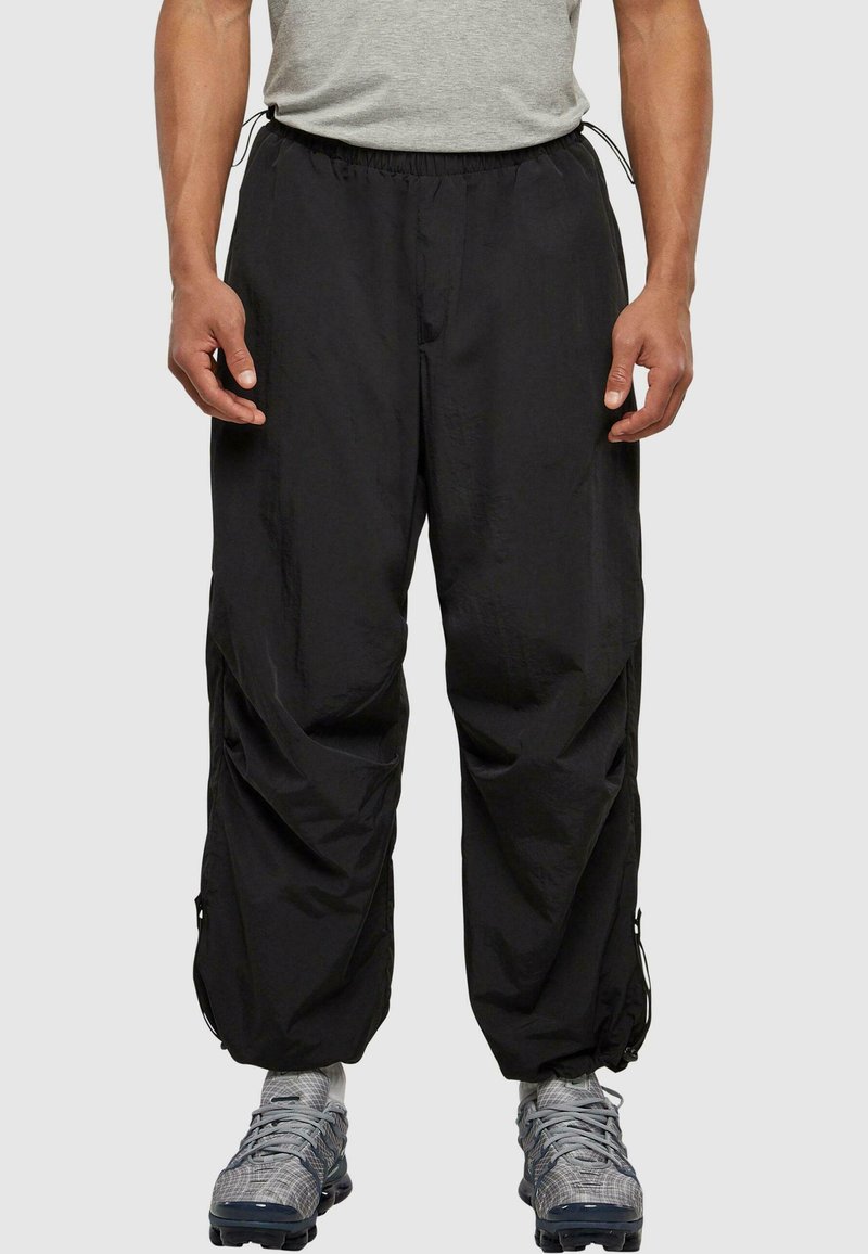 Sorte lette treningsbukser med elastisk midjebånd, sidelommer og justerbare mansjetter. Stoffet ser glatt og noe glansfullt ut.