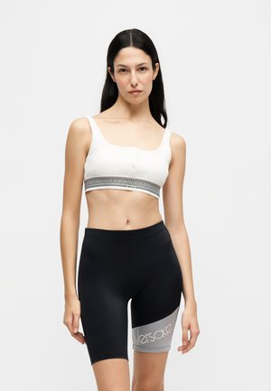 Femme portant un soutien-gorge de sport blanc avec un motif clé grecque et un short de cyclisme noir avec un panneau gris et le logo Versace.