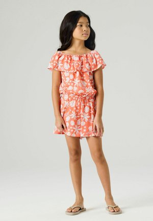 Pige, der står iført en koralrød off-shoulder kjole med hvid blomstret print og beige flip-flops, kigger til siden mod en ensartet baggrund.