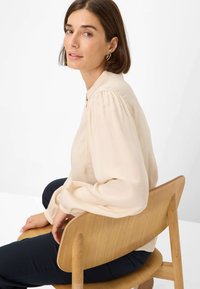 Beige blouse with puffed sleeves and a soft texture, buttoned at the collar, worn by a seated individual against a white background.
