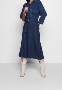 Vestido de denim con mangas largas, bolsillos frontales, un cinturón en la cintura y una falda acampanada. Combinado con botas de caña alta en color blanco sucio.