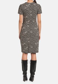 Žena v černých květinových šatech po kolena s krátkým rukávem a černých kozačkách na podpatku do půli stehen, stojící zády k fotoaparátu.