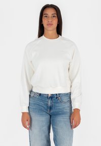 Felpa bianca con scollatura rotonda, polsini e orlo a costine. Abbinata a jeans denim blu, con una leggera scoloritura e chiusura a bottone.