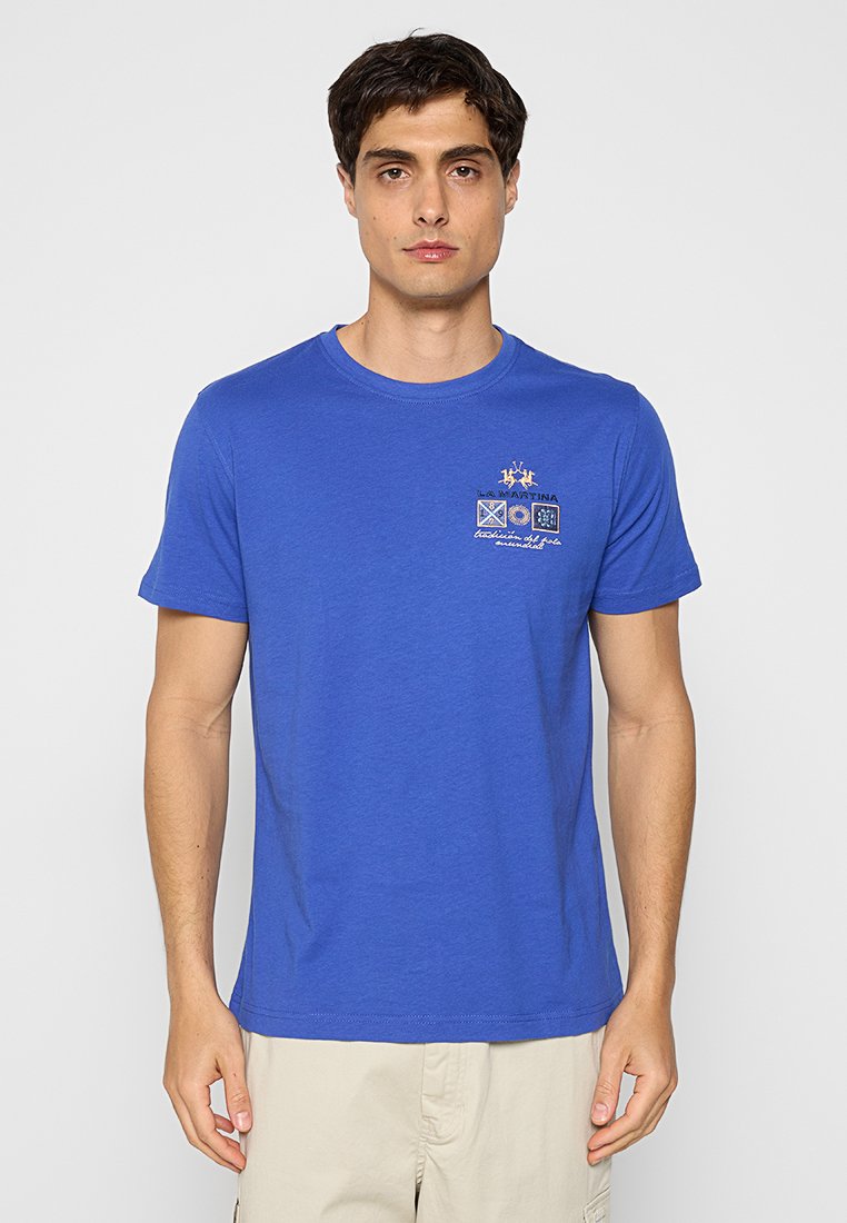 Mann, der ein blaues Kurzarmhemd mit kleinen gestickten Logos und beigen Hosen trägt, vor einem weißen Hintergrund steht.