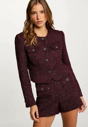 Giacca in tweed bordeaux con tasche frontali, finitura testurizzata e dettagli in bottone nero, abbinata a pantaloni shorts coordinati nello stesso materiale.