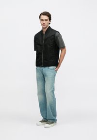 Schwarze gesteppte Weste mit Reißverschluss vorne und Stehkragen, kombiniert mit einem grauen Kurzarmshirt und hellblauen Jeans sowie grünen Turnschuhen.