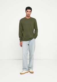 Olijfgroene langwerpige trui met een ronde hals, gecombineerd met lichtblauwe jeans en crèmekleurige sneakers met een gumrubberen zool. Eenvoudige, gestructureerde stof.