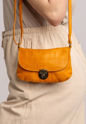 Vrouw draagt beige broek en top en draagt een kleine gele leren schoudertas met metalen sluiting ter hoogte van de heup.