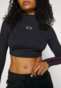 Musta lyhythihainen paita, jossa on korkea kaulus. Rinnassa on valkoinen logo ja hihoissa vaaleanpunaisia yksityiskohtia. Sujuva pinta.