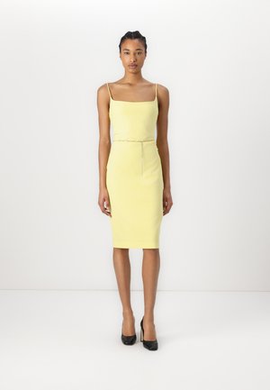 Yellow fitted dress with spaghetti straps, knee-length design, smooth texture, accentuated waist with a metallic chain detail, paired with black heels.