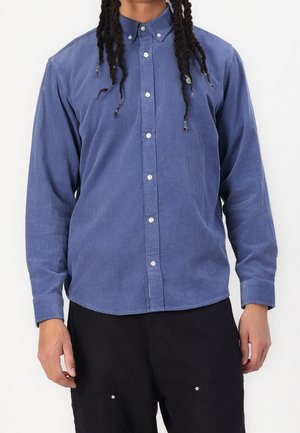Homme portant une chemise bleue à manches longues boutonnée avec un pantalon noir, debout devant un fond blanc uni.