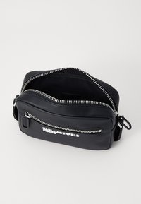 Borsa a tracolla nera in pelle con zip e logo argentati, dotata di una tasca frontale e texture liscia, aperta per mostrare l'interno.