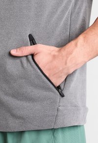 Poche grise zippée sur un vêtement en tissu texturé, avec une main insérée. La poche est équipée d'une fermeture éclair noire pour une fermeture sécurisée.