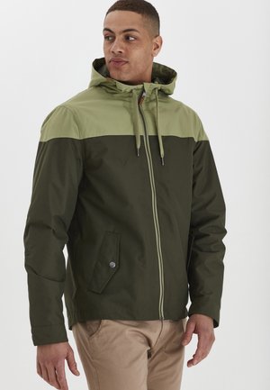 Veste colorée en vert foncé et vert clair. Fabriquée en tissu lisse avec une fermeture éclair, une capuche réglable et une poche latérale.