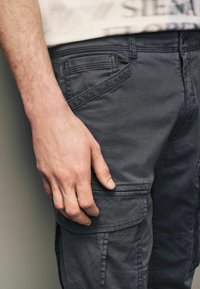 Grå cargo bukser lavet af bomuldsblanding, med sidelommer, subtil tekstur og tonet syning, vist med en hånd hvilende på stoffet.
