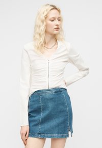 Lyshåret kvinde med bølget hår iført en hvid ruched bluse med lynlås og en blå denimmininederdel med frontslids, der kigger til siden.