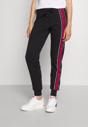 Persona con pantalones joggers negros con franjas laterales rosas con la inscripción "Love Moschino" y zapatillas blancas con detalles de corazones rojos, de pie contra un fondo liso.