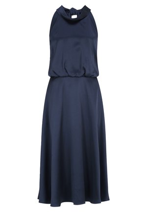 Femme portant une robe midi sans manches bleu marine à col halter, associée à des talons ouverts à bride cheville bleu pâle, debout sur un fond blanc.