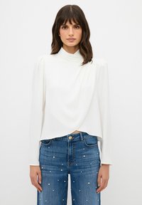 Top bianco a maniche lunghe con collo alto e design drappeggiato, abbinato a jeans in denim blu con dettagli in oro e taglio dritto.