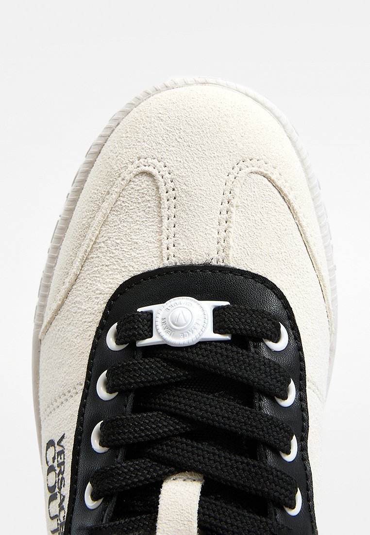 Zapatilla deportiva con parte superior de ante crema, ojales y cordones negros, con un detalle circular blanco en la lengüeta, suela de goma y logotipo metálico.