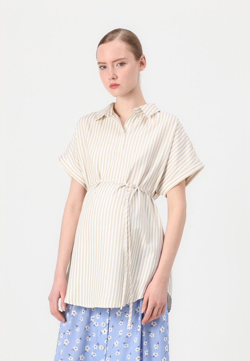 Camicia a righe beige chiaro con maniche corte, vestibilità rilassata e dettaglio a cintura. Abbinata a una gonna blu con motivo floreale.