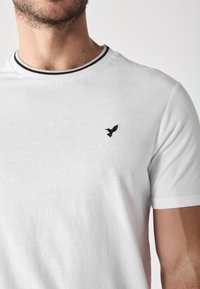 Weißes T-Shirt aus Baumwolle, mit einem schwarzen bestickten Vogel-Logo auf der linken Brust und einem schwarz gestreiften Kragen. Klassische Passform.