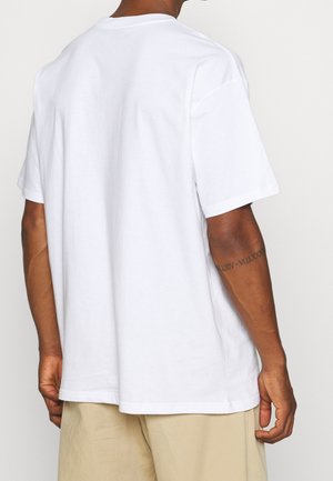 Mann trägt ein einfarbiges weißes kurzärmeliges T-Shirt und beige Shorts, von hinten gezeigt mit sichtbarem Tattoo am linken Unterarm.