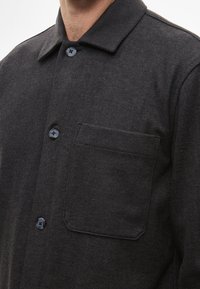Σκουρό γκρι πουκάμισο με υφή υφάσματος, διαθέτοντας γιακά, μπροστινή τσέπη και μαύρα κουμπιά, που αναδεικνύει έναν κλασικό σχεδιασμό.