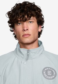 Lys grå jakke med høy krage, glidelås og rund logo-patch på venstre side. Jevnt materiale, avslappet passform og avrundede skuldre.