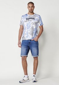 Balta un zila rakstota t-krekls ar "KORÖSHI Brand" uzrakstu, apvienots ar ziliem džinsu īsajiem šortiem un pelēkām un baltām sporta apaviem.