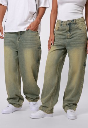 Baggy denim jeans with a faded green and yellow wash, featuring five pockets, belt loops, and embellished stitching. Worn with white sneakers.