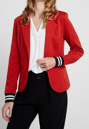 Femme portant un blazer rouge avec des poignets rayés noirs et blancs sur un chemisier blanc à col en V et un pantalon noir, posant avec une main près du blazer.