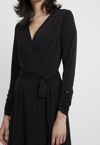 Svart omslagsklänning med långa ärmar, med v-ringning, knytbälte, rynkade ärmdetaljer och slät tygstruktur.