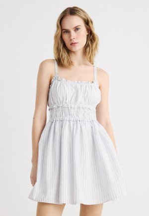 Junge Frau mit schulterlangem blondem Haar, die ein hellgraues ärmelloses Sommerkleid mit elastischem Raffungseinsatz und dezenten vertikalen Streifen trägt.