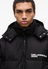 Sort pufferjakke med høj krave, lynlås og trykknapdetaljer. Har et synligt logo: "KARL LAGERFELD JEANS." Glat tekstur.