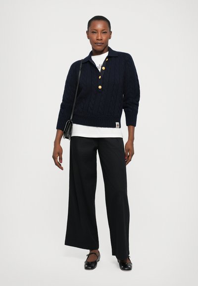 Pull col roulé en maille torsadée marine avec un col et des boutons dorés, superposé à une chemise blanche. Associé à un pantalon noir à jambes larges et des ballerines.