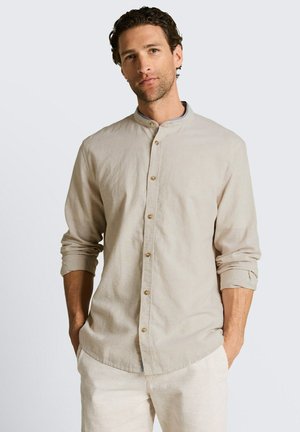 Homme aux cheveux bouclés portant une chemise beige à col mandarin boutonnée et un pantalon clair, mains dans les poches, fond neutre.