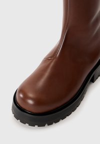 Rjavi usnjeni škorenj z zaobljenim nosom in debelim črnim zatičem na belem ozadju, ki prikazuje sprednje in stranske podrobnosti.