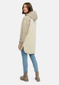 Beige hættetrøje med quiltede ærmer og en blød, fluffy krop. Har et logo på ærmet og slidser i siden. Bæres over blå jeans og brune støvler.