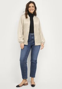 Beige satijnen bomberjack met elastische manchetten, gecombineerd met donkere denim jeans en twee-tint flats. Zwarte coltrui eronder.