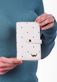 Bílá peněženka s černými puntíky, hladká textura, zacvakávací uzávěr a zlatý akcent s logem na spodní straně. Má obdélníkový tvar.